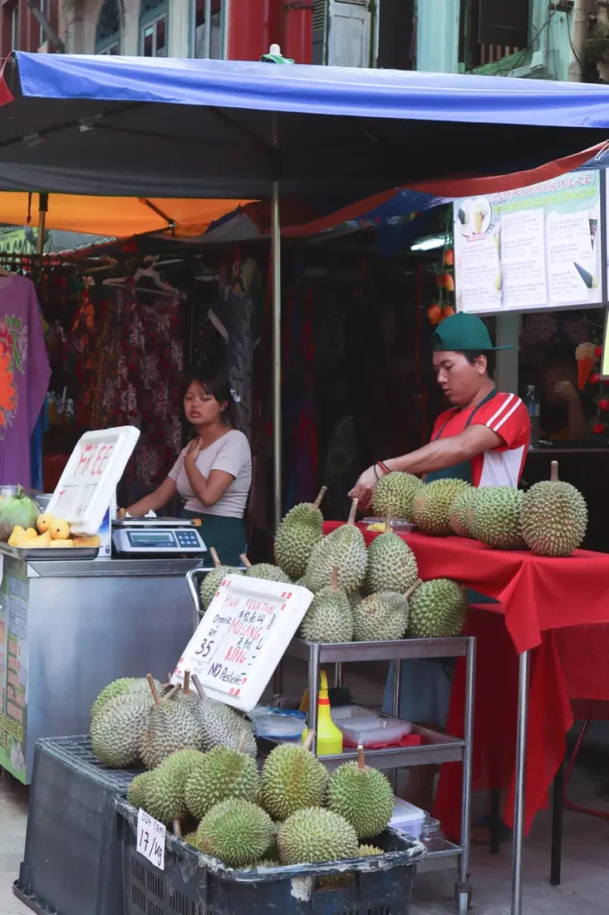 A street vendor in Kuala Lumpur preparing durians at a market stall, surrounded by fresh tropical fruits and colorful awnings.