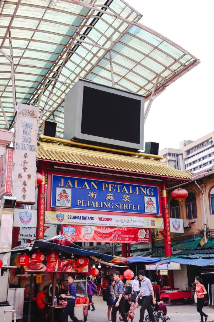 Entrance to Petaling Street in Kuala Lumpur, adorned with bright signs and lanterns, with bustling street vendors and visitors.