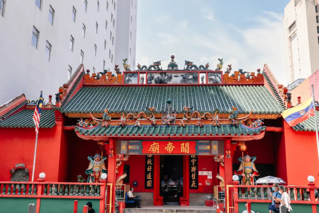 Vibrant red Guan Di Temple in Kuala Lumpur with intricate carvings and statues, contrasting against a modern city backdrop