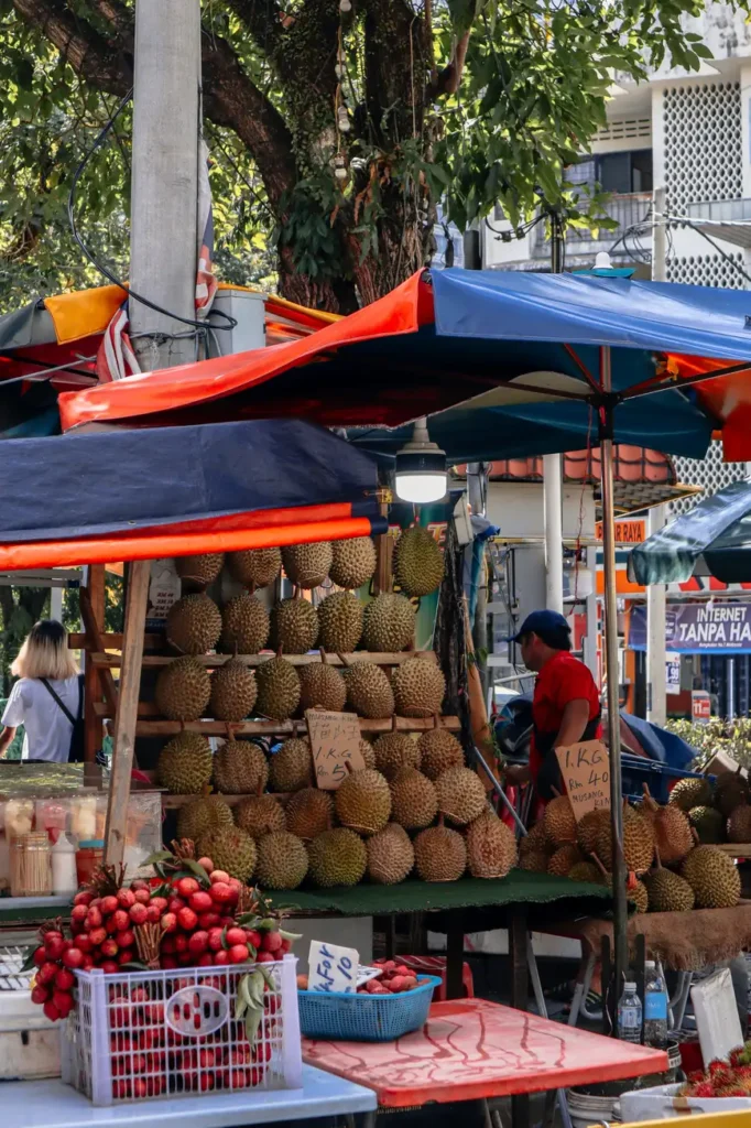 A bustling street market in Kuala Lumpur featuring neatly stacked durians and other tropical fruits with vibrant market stalls in the background.