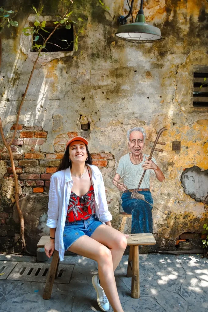 Me smilling seated next to a painted mural of an elderly musician playing a traditional instrument at Kwai Chai Hong in Kuala Lumpur