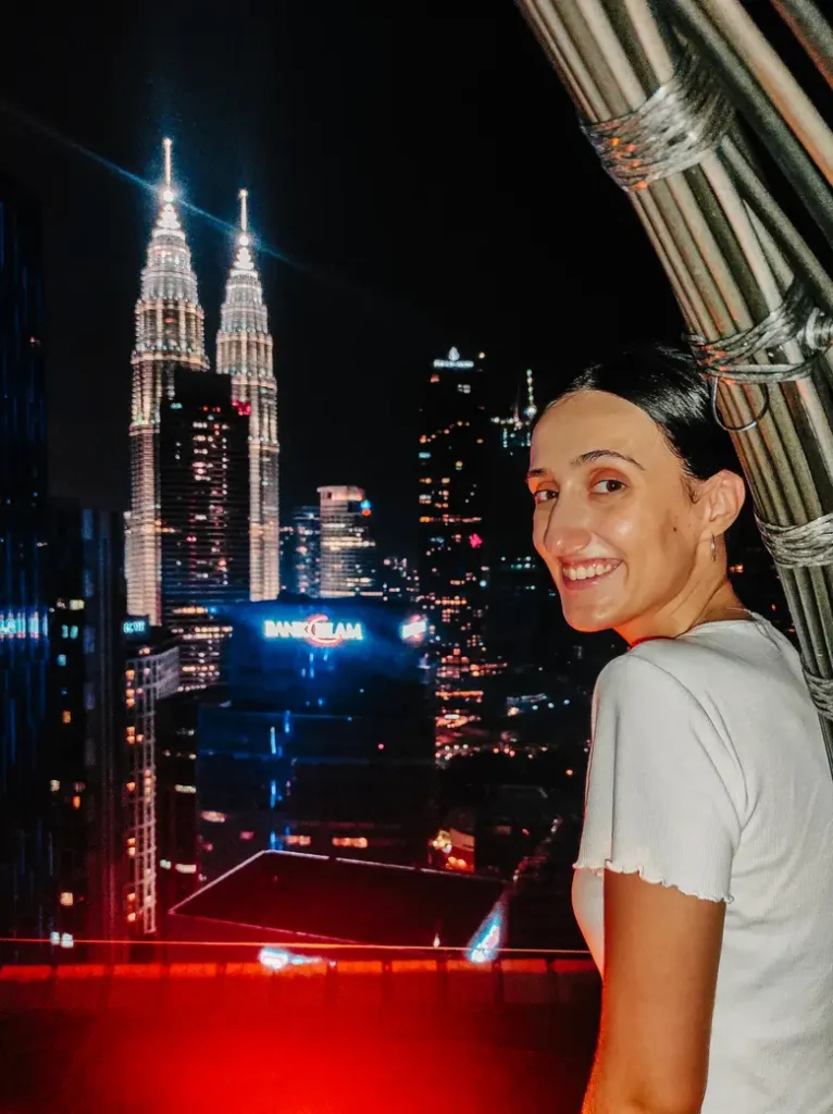 Smiling woman at Helipad Rooftop Bar with the iconic Petronas Twin Towers illuminated in the background, Kuala Lumpur at night