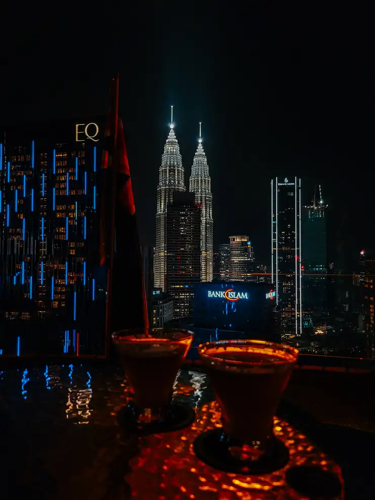 A nighttime view of Kuala Lumpur's skyline featuring the illuminated Petronas Twin Towers, seen from a rooftop bar with drinks in the foreground.