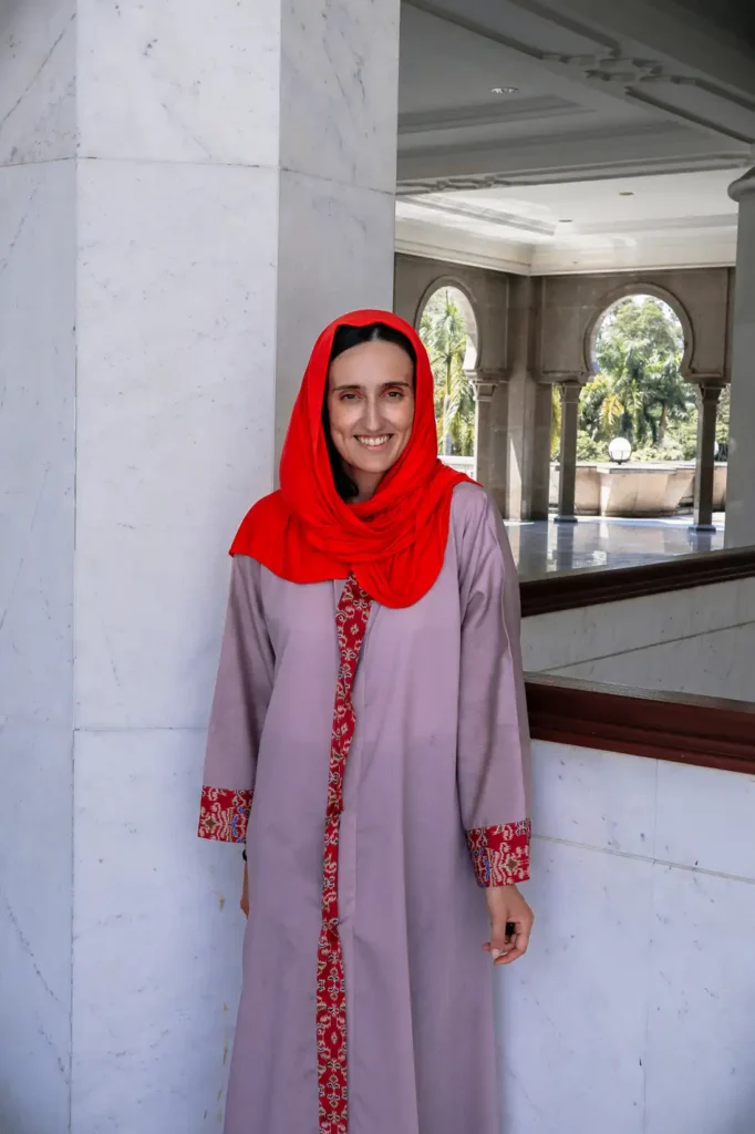 A person wearing a traditional robe and red headscarf standing inside the Wilayah Mosque, showcasing the grand architecture with arched windows and marble columns.