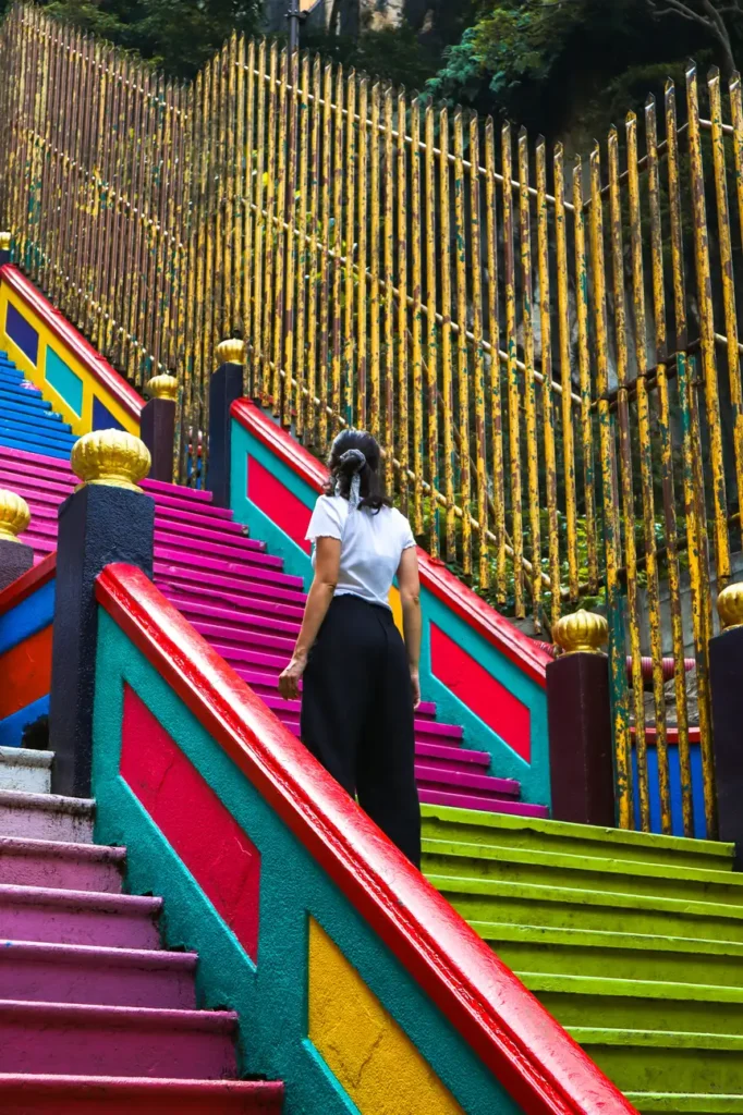 A person walking up the vibrant, multicoloured staircase of the Batu Caves, with golden railings and lush greenery in the background.