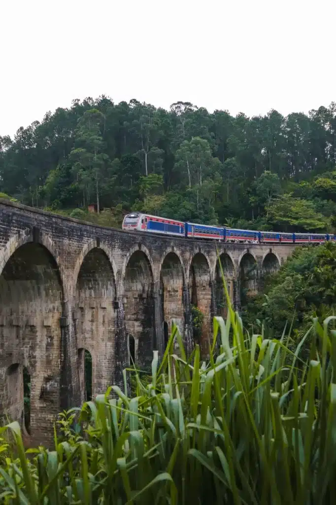 A scenic view of the iconic Nine Arches Bridge in Ella, Sri Lanka, with a train passing over its stone arches, surrounded by lush greenery and forested hills