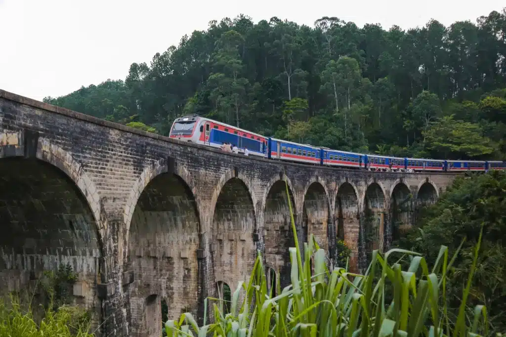 A vibrant train crossing the iconic Nine Arches Bridge in Ella, Sri Lanka, with its historic stone arches framed by lush green foliage