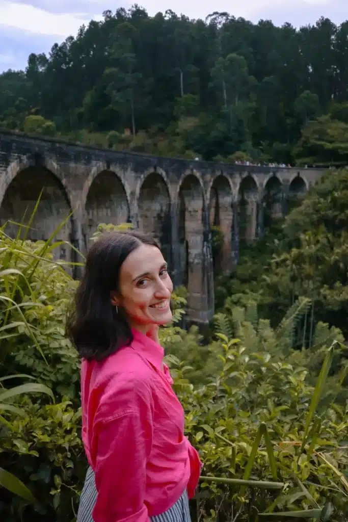 A smiling woman in a bright pink shirt standing near the famous Nine Arches Bridge in Ella, Sri Lanka, with the lush green landscape and the arched structure in the background