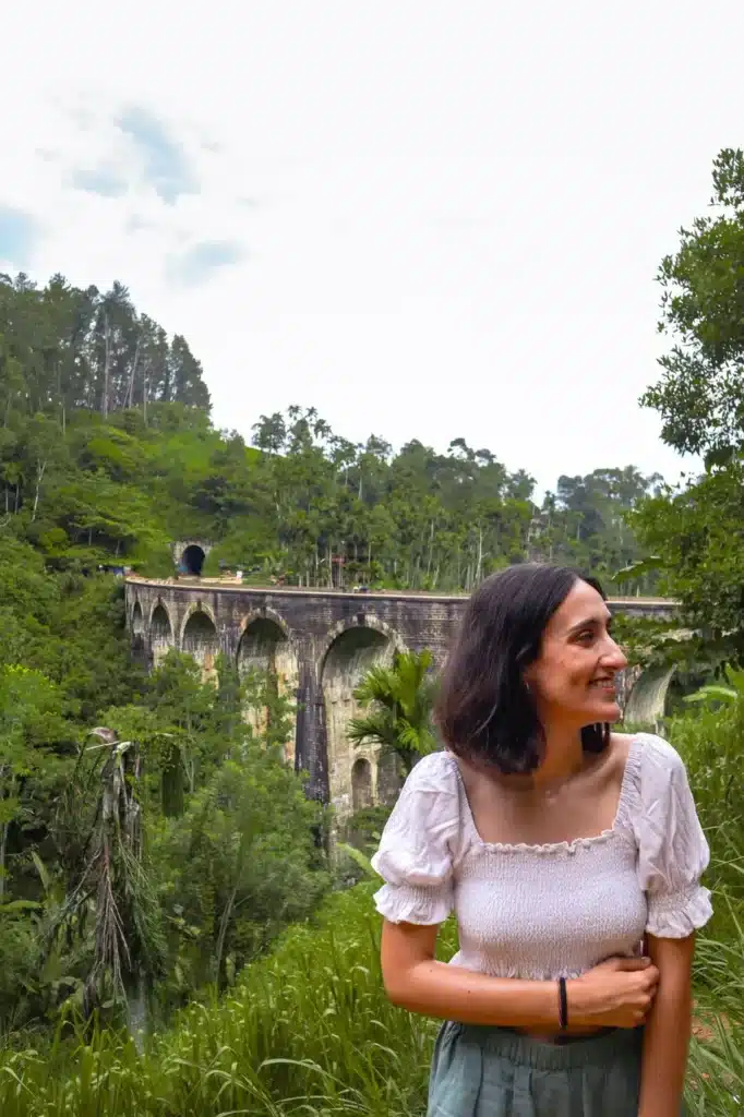 A cheerful woman wearing a white top and standing near the iconic Nine Arches Bridge in Ella, Sri Lanka, surrounded by vibrant greenery