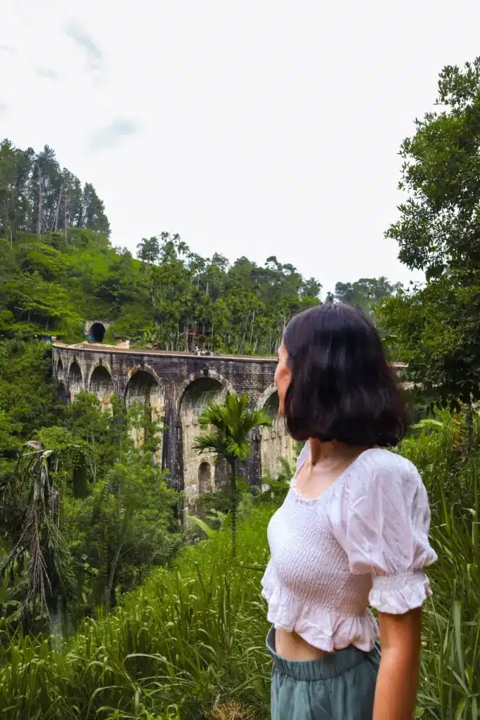 A woman in a white top admiring the Nine Arches Bridge in Ella, Sri Lanka, surrounded by vibrant greenery and tropical trees