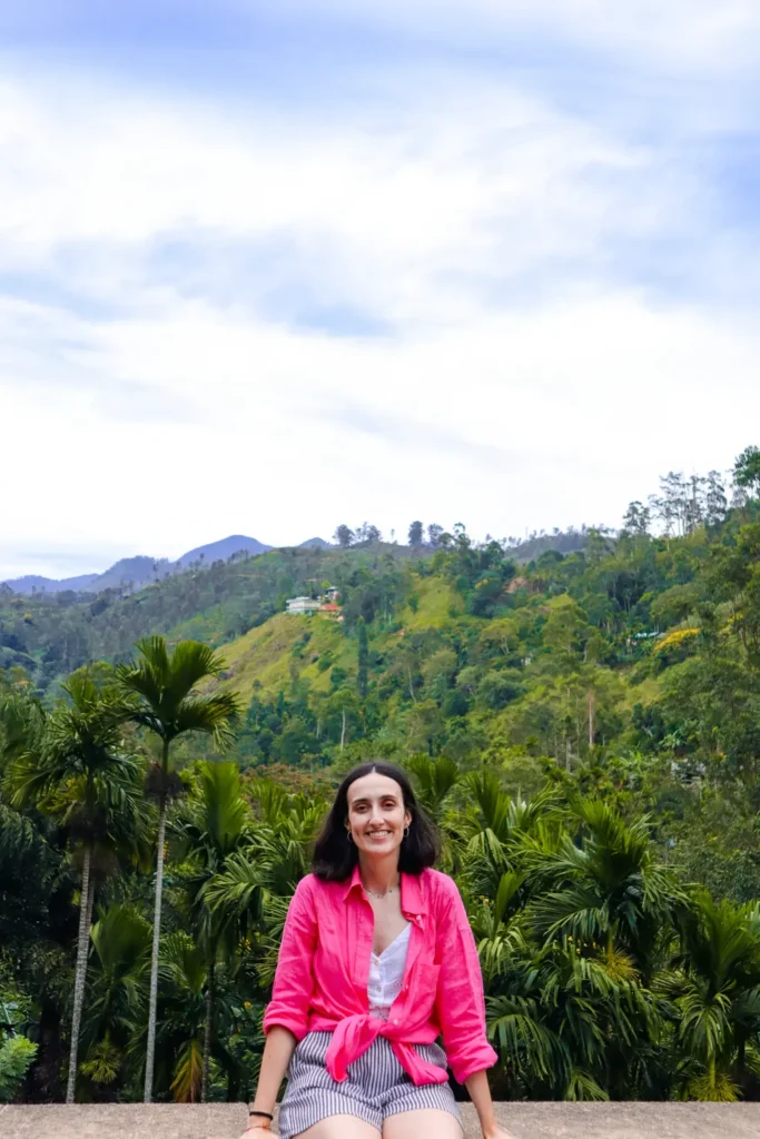 A smiling woman in a pink shirt sitting against a lush green hillside backdrop in Ella, Sri Lanka, with tall palm trees and scenic mountain views