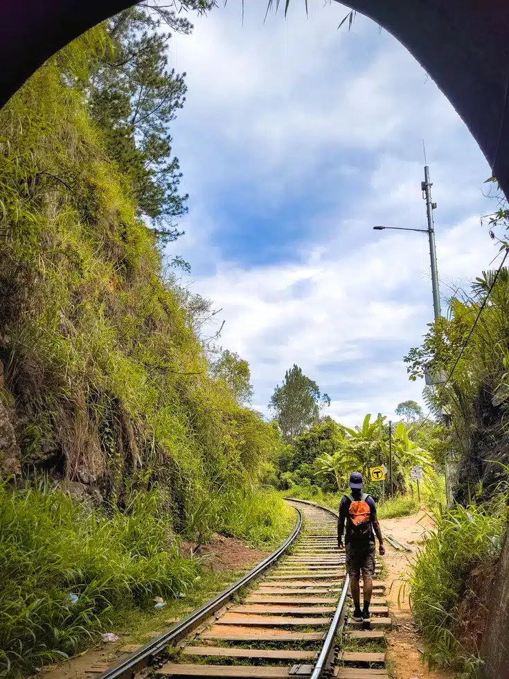 A hiker walking along the railway tracks leading to the Nine Arches Bridge in Ella, Sri Lanka, framed by lush vegetation and a bright blue sky