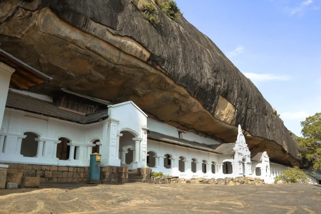 The Dambulla Cave Temple complex, featuring a line of white buildings built into a massive rock face under a towering cliff. A popular day trip destination from Kandy and a must-visit cultural site in Sri Lanka.