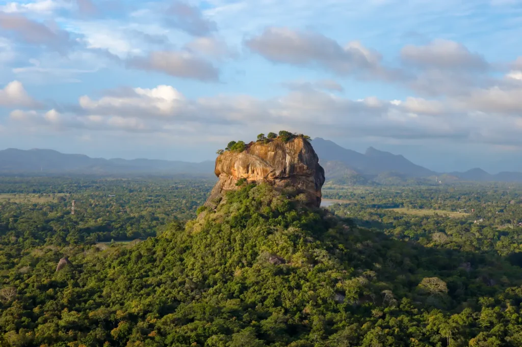 A majestic view of Sigiriya, the Lion Rock Fortress, surrounded by dense greenery under a clear sky. This iconic UNESCO World Heritage Site is a memorable day trip from Kandy and a top cultural attraction in Sri Lanka.