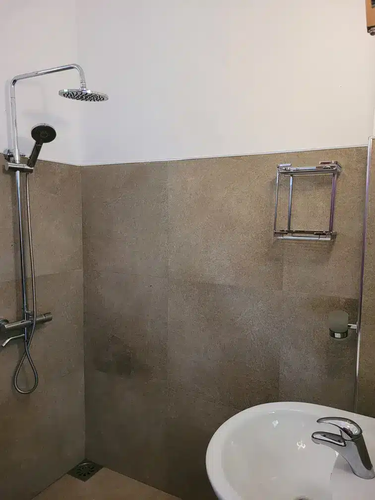 A minimalist bathroom with beige tiled walls, a modern rainfall shower, and a sleek white sink, showcasing the amenities of hotels in Ella, Sri Lanka