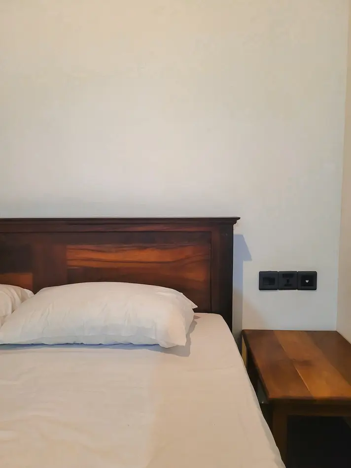 A simple wooden bed with white linens and a matching wooden bedside table in a cozy hotel room. This image highlights the comfortable accommodations available in hotels in Kandy.