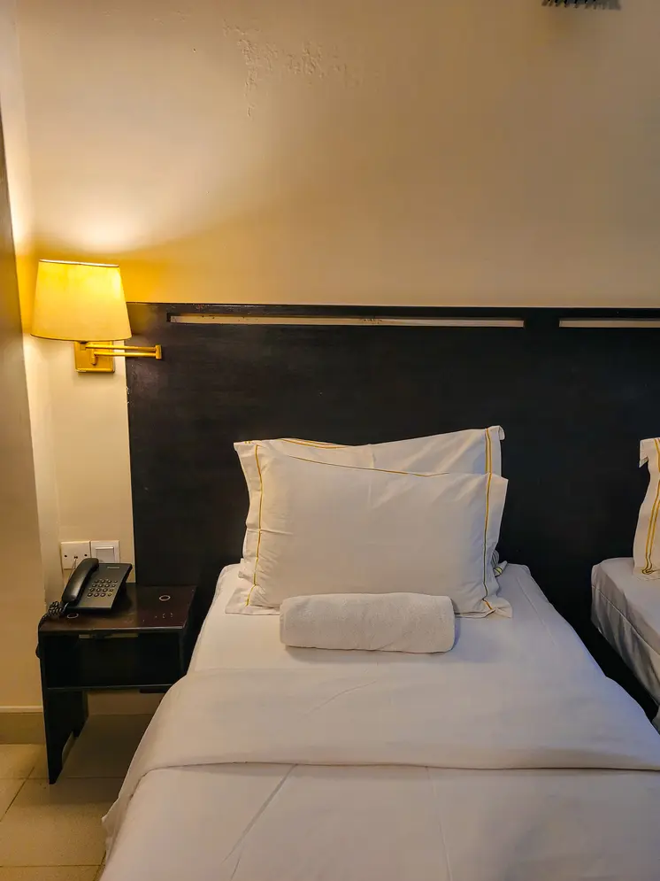 A neatly made bed with soft white pillows and a rolled towel, accompanied by a bedside lamp casting a warm glow. This showcases the inviting and modern rooms found in Kandy hotels.