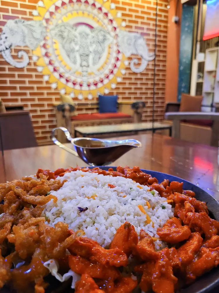 A colorful plate of rice with spiced chicken and crispy vegetables, set against a vibrant restaurant decor with an elephant mural. This highlights the flavorful dining experiences in Kandy.