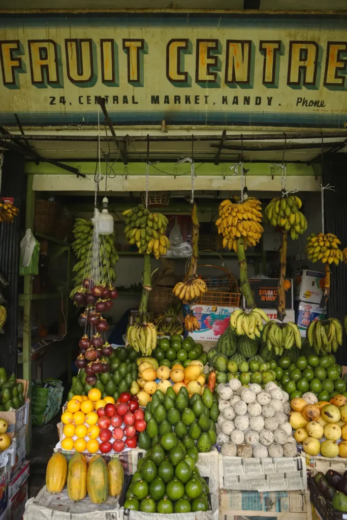 The entrance to a fruit shop at Kandy Central Market, displaying neatly arranged tropical fruits like bananas, avocados, and dragon fruit. Visiting this market is a popular activity for those seeking fresh produce and a lively atmosphere in Kandy.