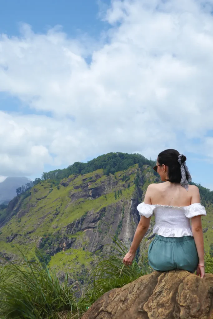 A woman sits on a rock at Little Adam's Peak in Ella, Sri Lanka, enjoying the lush green mountain views under a bright blue sky. Visiting Little Adam's Peak is one of the best things to do in Ella, offering stunning scenery and a peaceful hiking experience.