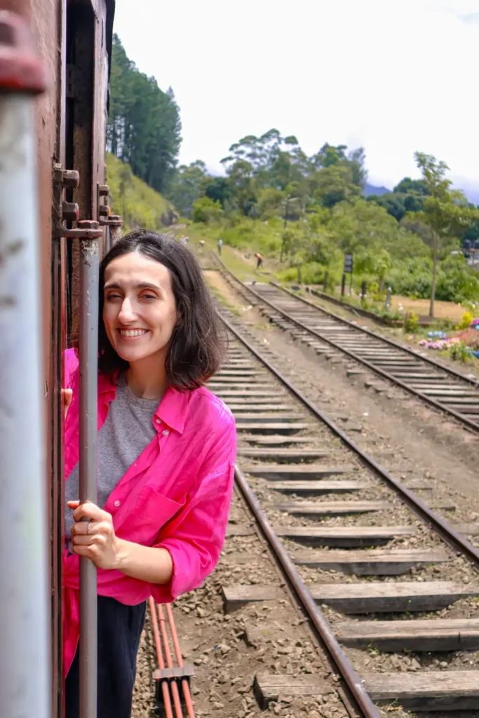 A woman in a bright pink shirt smiles while leaning out of a train door, with railway tracks and greenery visible in the background on a clear day