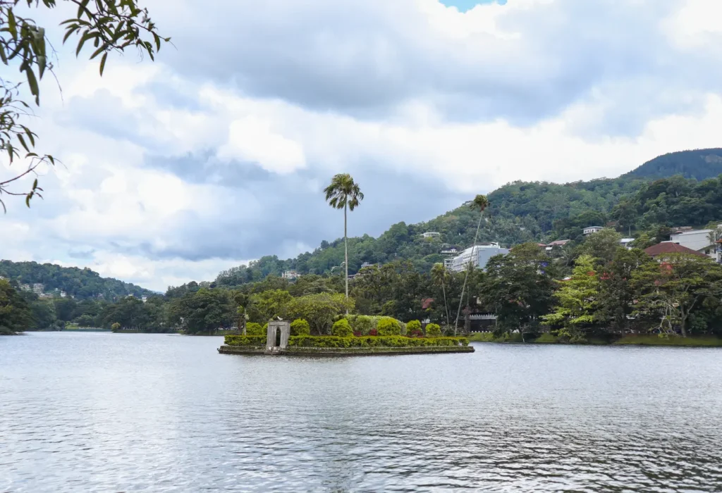 A scenic view of Kandy Lake, showcasing a small green island with palm trees and a stone structure at its center. The surrounding hills and lush vegetation reflect on the water under a partly cloudy sky.