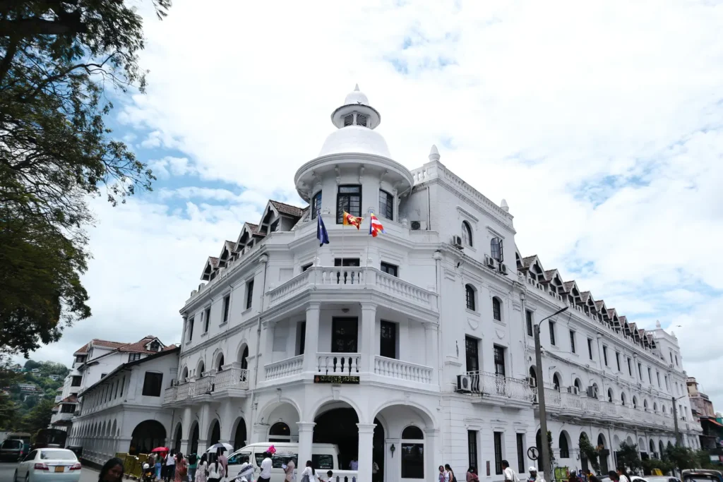 : The iconic white facade of the Queen’s Hotel in Kandy, Sri Lanka, with flags fluttering on its balcony. The historic colonial-style building stands out under a bright blue sky, surrounded by bustling streets.