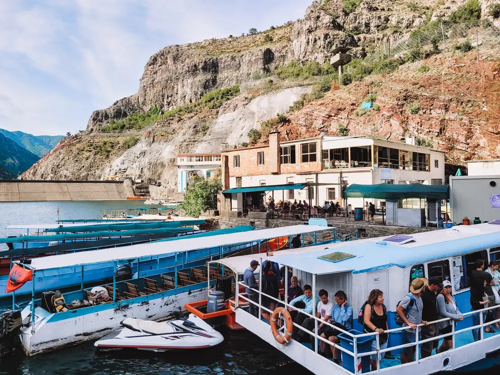 A busy ferry dock on Lake Koman with vibrant boats and passengers against a backdrop of rugged cliffs and small buildings, showcasing the lively hub for lake tours.