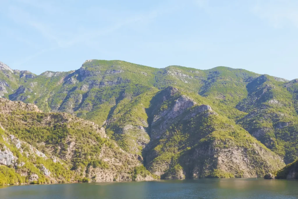 A tranquil view of Lake Koman framed by steep, forested cliffs and serene blue waters, capturing the lake’s natural beauty and peaceful atmosphere