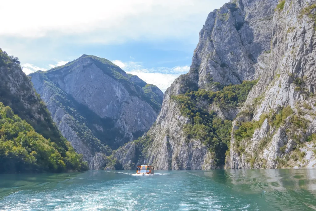 A tranquil view of Lake Koman framed by steep, forested cliffs and serene blue waters, capturing the lake’s natural beauty and peaceful atmosphere