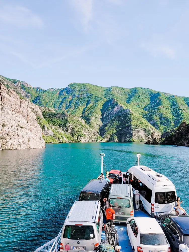 Vehicles and passengers aboard a ferry crossing Lake Koman, surrounded by dramatic green hills and clear blue waters, showcasing the unique transport experience in this Albanian destination