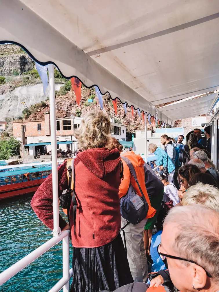 Passengers aboard a covered ferry near Lake Koman’s dock, with views of red and blue boats and rugged cliffs in the background, highlighting the vibrant ferry hub.