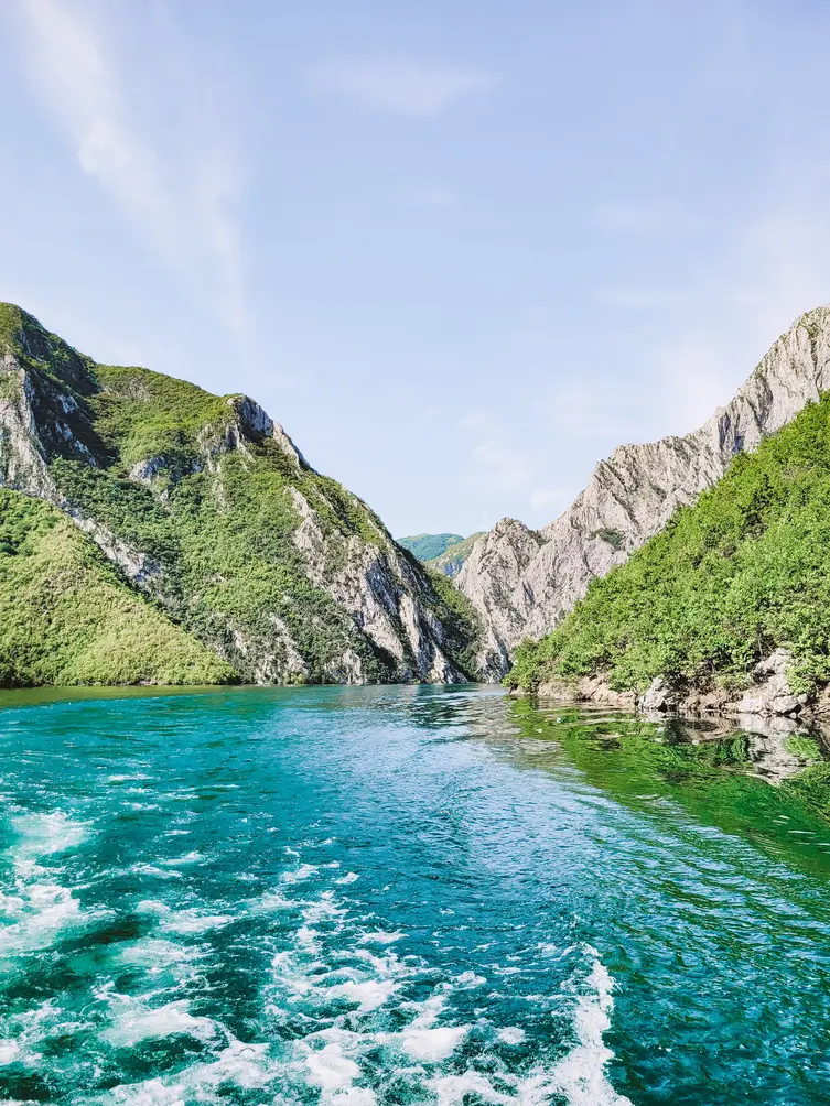 A tranquil view of Lake Koman framed by steep, forested cliffs and serene blue waters, capturing the lake’s natural beauty and peaceful atmosphere