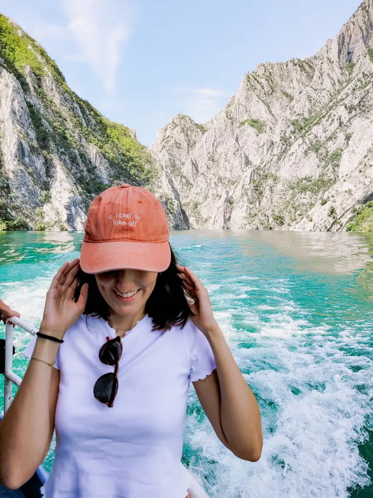 A smiling woman wearing a peach cap with the phrase 'a ticket to take off,' standing on a ferry on Lake Koman, surrounded by turquoise waters and dramatic rocky cliffs, highlighting the lake’s scenic and adventurous appeal.