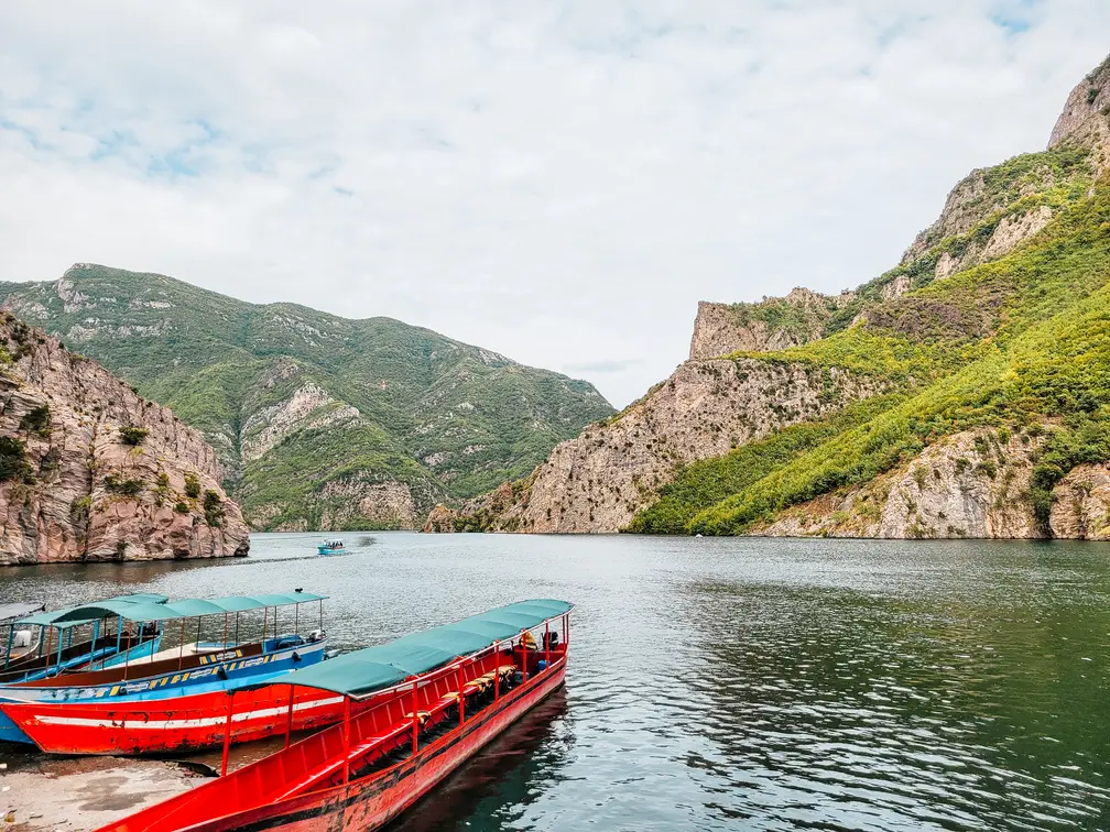 Brightly colored red and blue boats docked along the calm waters of Lake Koman, framed by towering cliffs and green hills, offering an idyllic view of the lake's boating area