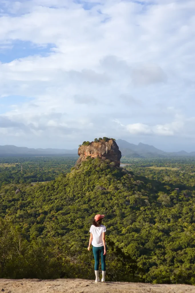 A hiker in a white shirt and pink cap stands on Pidurangala Rock, admiring the stunning view of Lion Rock rising above the lush green forest