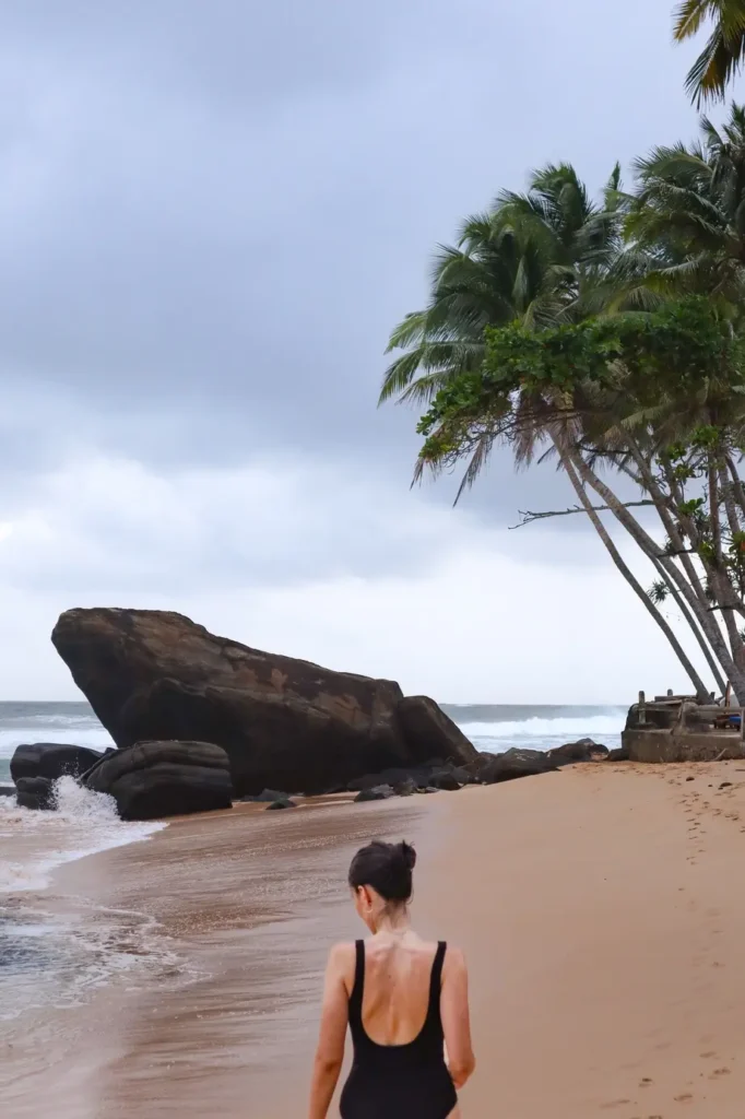Mariana strolls along an empty shoreline toward a dramatic boulder, a serene experience perfect for any Mirissa itinerary.