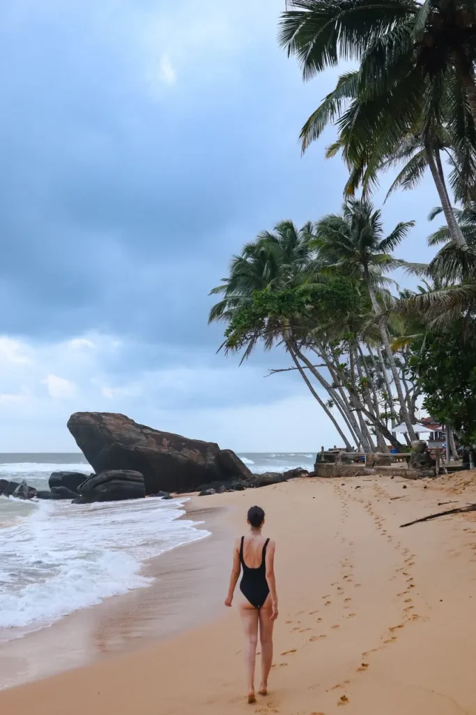 Barefoot Mariana walks along the wet sand, leaving footprints toward the iconic boulder and leaning palms—an unmissable stop on your list of places to visit in Mirissa.