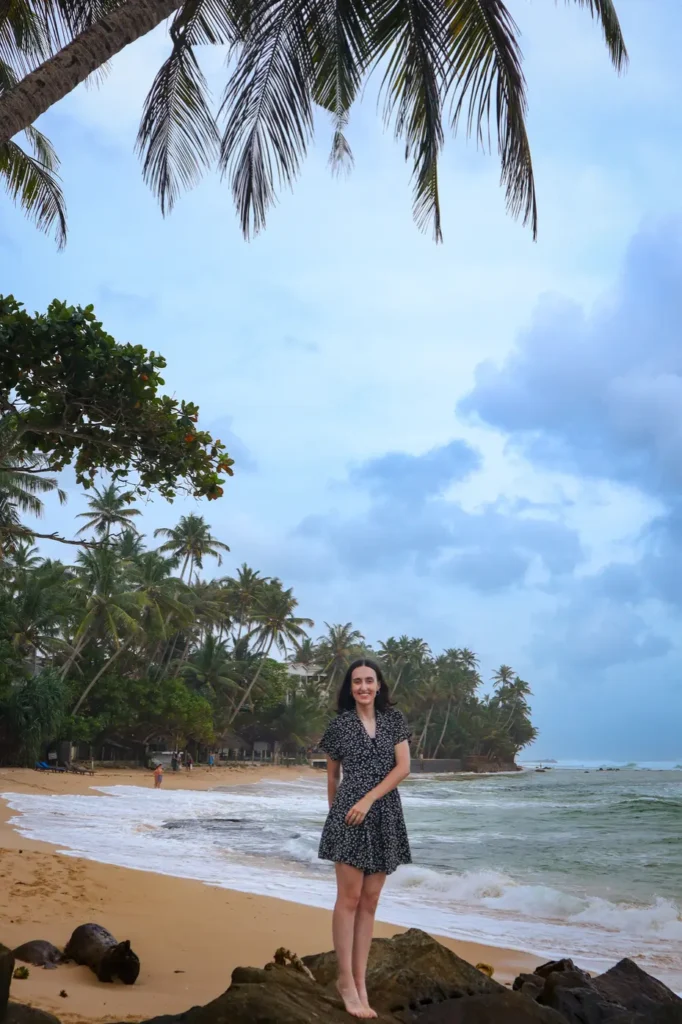 Mariana poses on rocks at the edge of a tropical beach, backed by lush coconut trees and waves—definitely one of the top things to do in Mirissa.