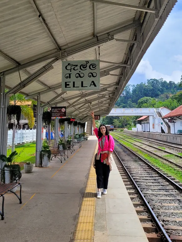 Mariana stands on the platform at Ella train station, pointing up at the station sign with “ELLA” written in both English and Sinhala. It’s a popular departure point for the scenic kandy to ella train journey through Sri Lanka’s hill country.