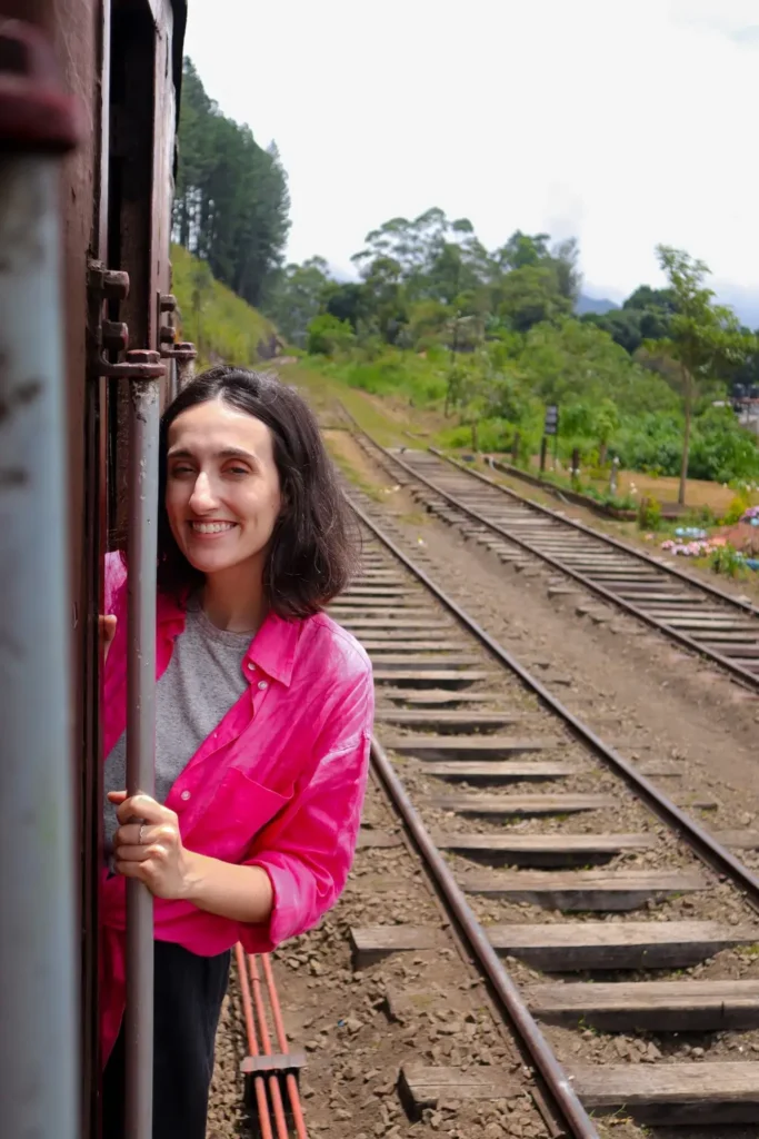 Close-up of smiling Mariana hanging out of a moving train, surrounded by green hills and railway tracks. The ella to kandy train offers unforgettable moments like these, where passengers ride with the wind in their hair.