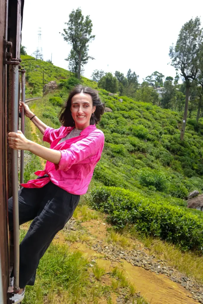 Mariana in a pink shirt holds onto the train rail while passing through vibrant green tea plantations on the kandy to ella train. The open-door ride makes it easy to soak in the scenery of Sri Lanka’s lush countryside.