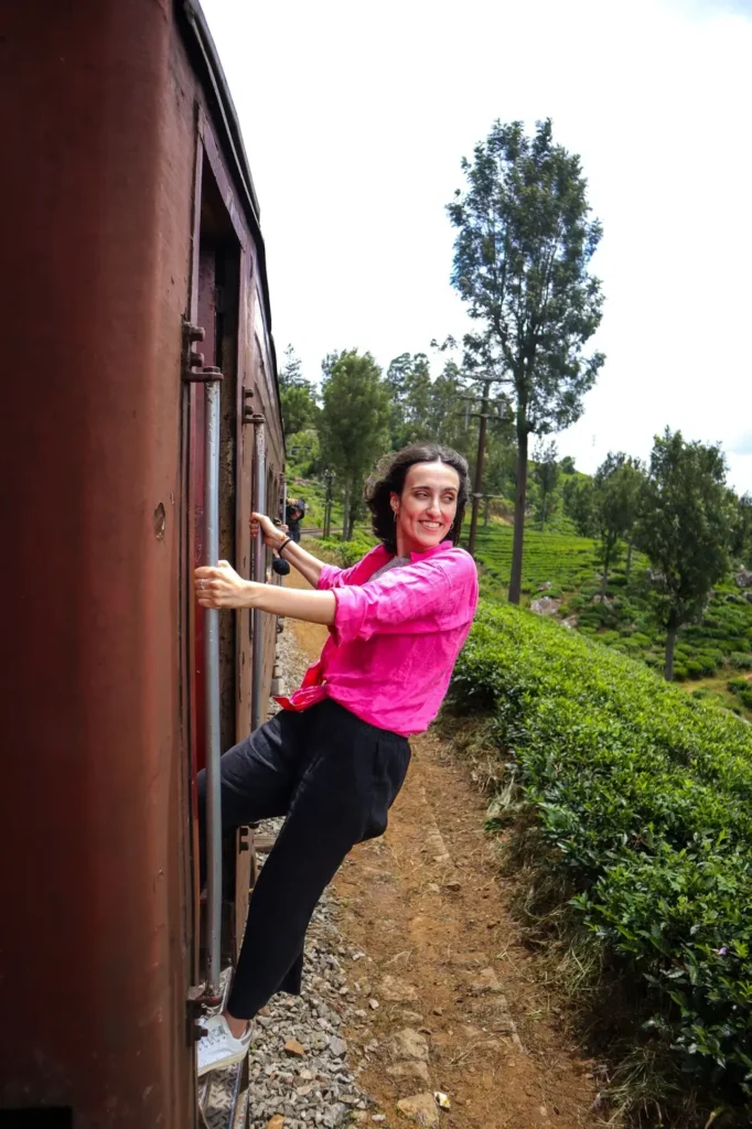 Riding the ella to kandy train, smiling Mariana leans out of a red carriage surrounded by dense greenery and tea fields. The train snakes through the landscape, making every turn a photo-worthy moment.