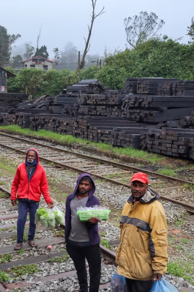 Group of local men, some holding baskets and plastic bags of produce, walk beside the tracks near the kandy to ella train route. Behind them, stacks of railway sleepers and misty hills set a moody backdrop.