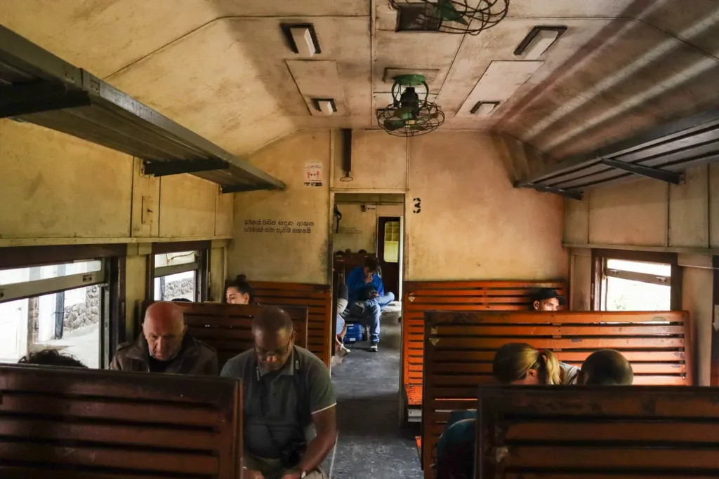 Interior of a third-class train car on the ella to kandy route, with wooden benches, ceiling fans, and a mix of passengers. The vintage setting reflects the charm and authenticity of train travel in Sri Lanka.