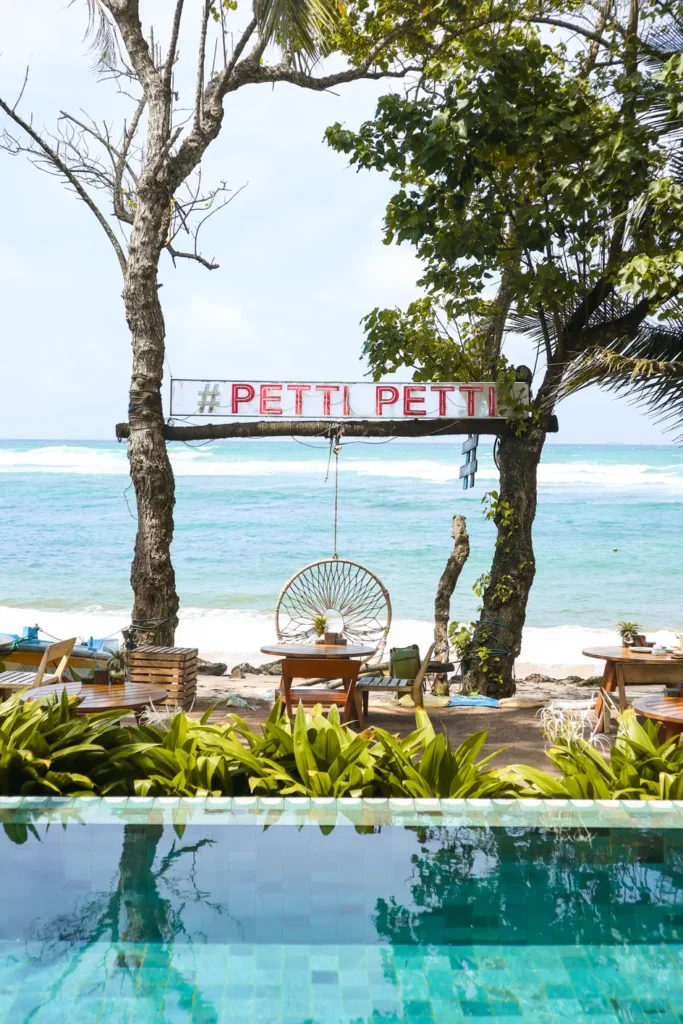 Outdoor beachfront seating at Petti Petti Beach Club with wooden tables, a circular hanging chair, tropical plants, and a turquoise pool in the foreground. A sign reading "#PETTI PETTI" hangs between two trees with the ocean waves and blue sky in the background.