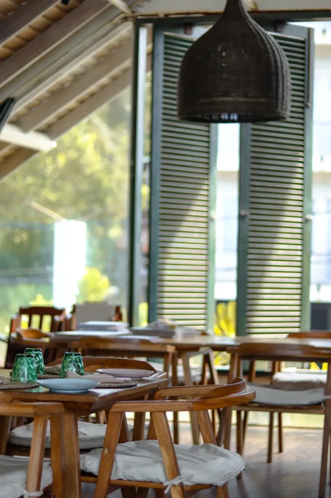 Cozy indoor seating area at Petti Petti Beach Club featuring wooden tables and chairs with white cushions, green-tinted glasses set on placemats, and sunlight streaming through open green shutters.