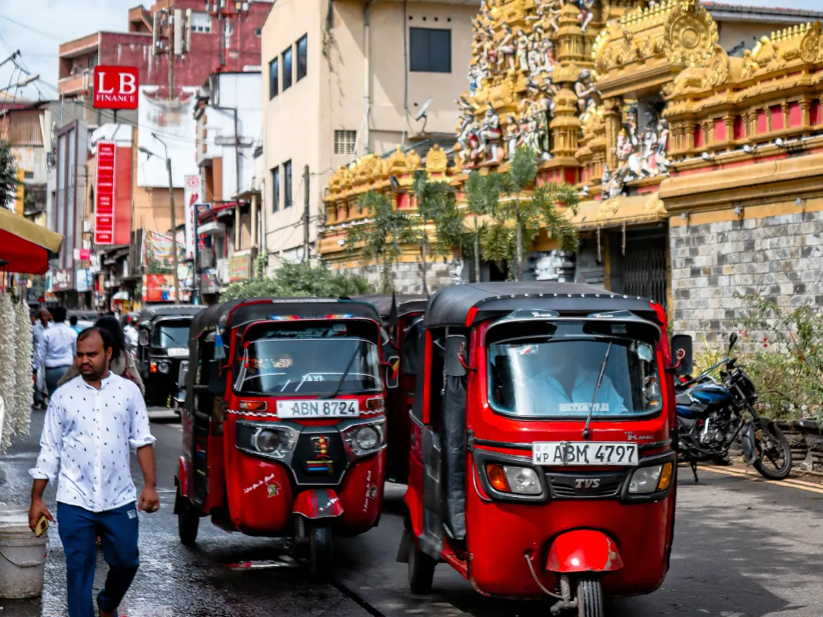 Busy street scene in Sri Lanka with bright red tuk-tuks navigating past pedestrians and a golden Hindu temple adorned with intricate carvings in the background. This vibrant local moment reflects the energy and culture you can expect when planning a trip to Sri Lanka.