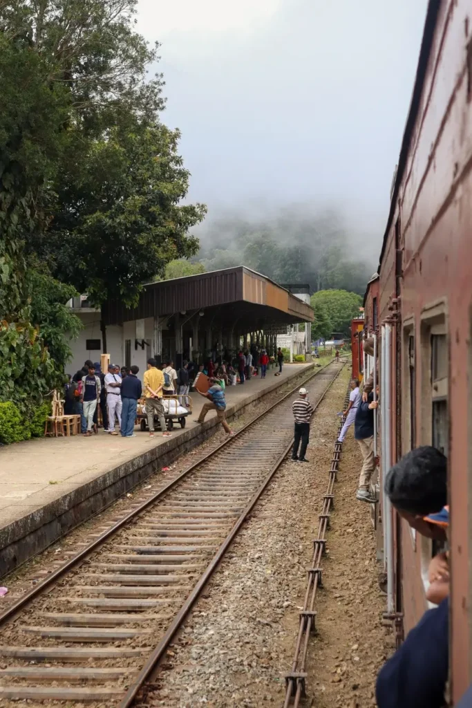 Passengers and vendors gather at a misty hillside station as the kandy to ella train pulls in. Locals prepare carts and luggage while travelers peek out of the train’s windows.