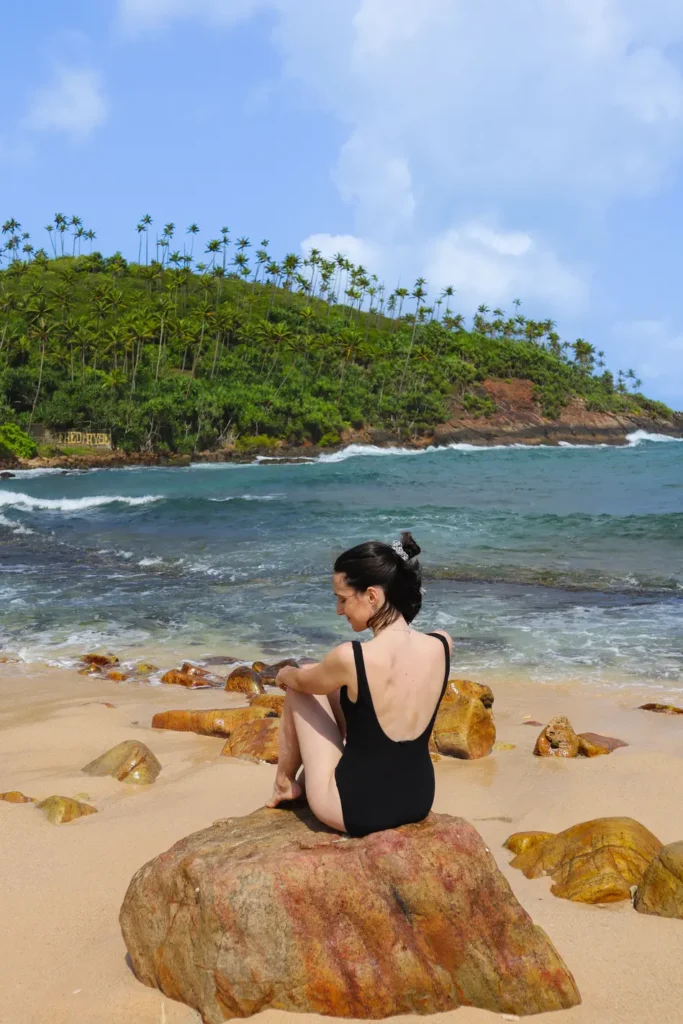 Mariana in a black swimsuit sits on a large rock by the shore, with palm-covered hills and turquoise waves in the background—showcasing the serene beauty of Secret Beach, one of the top things to do in Mirissa.